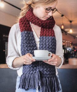 Suzette stitch long crochet scarf in burgundy and blue with tassel fringe, worn in a coffee shop