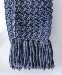Suzette stitch crochet scarf in blue with tassel fringe hanging against white wall